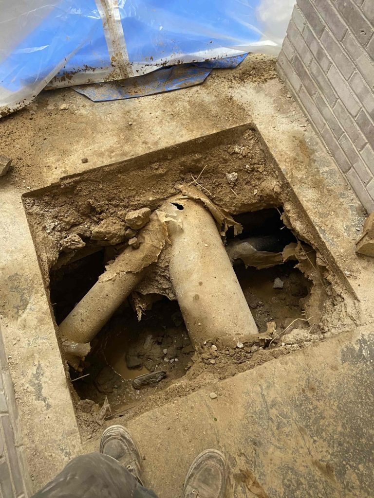 A top-down view of a square hole cut into a concrete floor, exposing underground dirt and two large, weathered utility pipes crossing each other. A worker's boots are visible at the bottom edge of the frame, with a brick wall and plastic dust containment sheeting nearby in the background.
