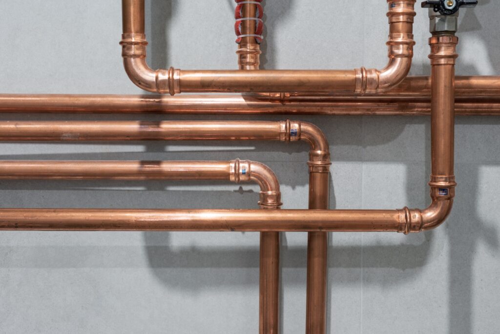 A detailed view of a modern copper plumbing installation against a flat, light grey wall. The shiny pipes are arranged in a clean grid of horizontal and vertical runs, utilizing press-fit elbow and T-joints, some featuring small blue indicator marks. At the top, one pipe has red and white wrapping, and another features a black shut-off valve handle. Bright lighting casts sharp shadows of the pipe network onto the wall behind it.