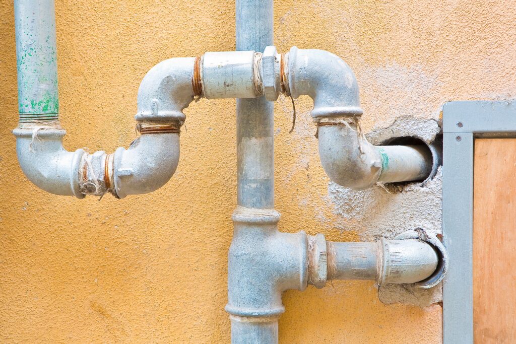An exterior view of aging galvanized steel plumbing. A vertical pipe features a T-junction, while a secondary pipe routes from the top left using multiple elbow fittings. The threaded joints show signs of corrosion and fraying plumber's hemp. On the right, two pipe segments enter the building through roughly chipped holes in a textured orange wall.