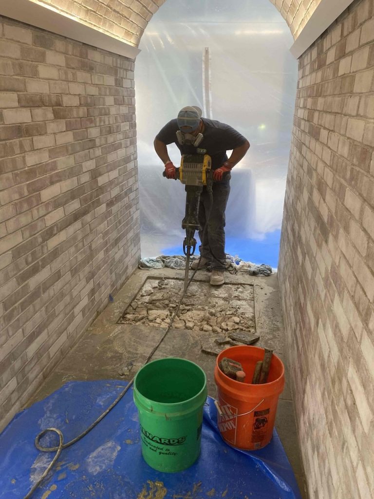 A Eco technician worker wearing a respirator mask uses a jackhammer to break up a section of concrete flooring inside a narrow, arched brick hallway. In the foreground, a green bucket and an orange bucket sit on a blue tarp, while the background is sealed off with clear plastic sheeting for dust control.